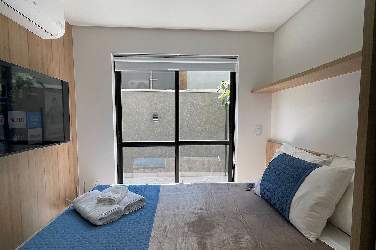 A hotel room with a bed, a TV, a window, and a ceiling light.