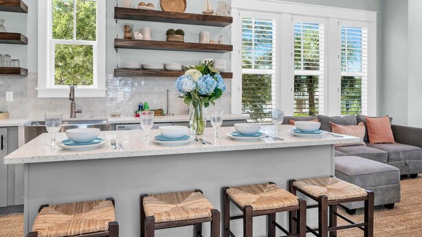 A modern kitchen with a central island, wooden shelves, and a dining area with a grey sofa and wicker stools. - 🦩Book Me Baby! Seaglass in Santa Rosa Beach  ~ Golf Cart Included🦩 (Santa Rosa Beach)