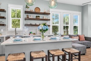 A modern kitchen with a central island, wooden shelves, and a dining area with a grey sofa and wicker stools.