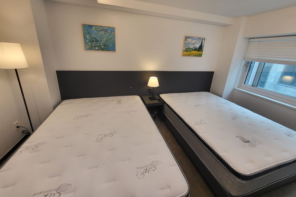 Quarto de hotel com duas camas, mesa de cabeceira com abajur, janela com persianas e dois quadros na parede.