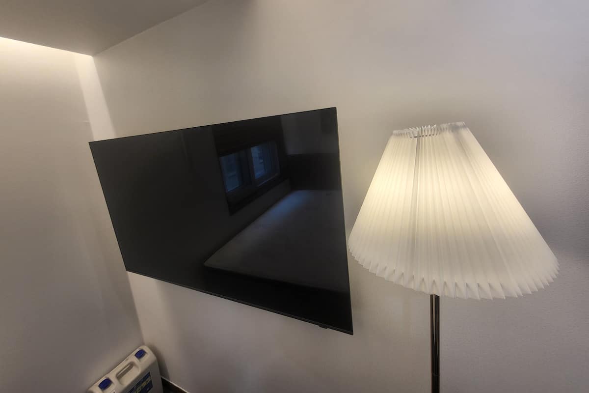 Televisão de tela plana presa na parede, um abajur de chão e uma pequena janela com persianas.