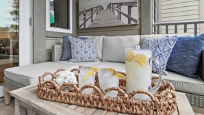 Un plateau en osier contenant des boissons et une carafe, posé sur une table, un canapé agrémenté de coussins à motifs et un tableau représentant une jetée.