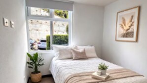 Een slaapkamer met een bed, een raam, een plant en een lijst met een afbeelding aan de muur.