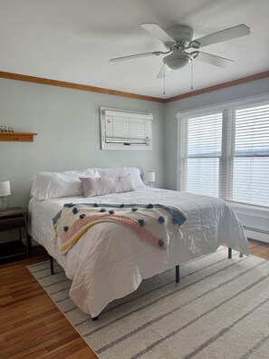 Une chambre à coucher comprenant un lit, un ventilateur de plafond, une fenêtre avec des stores, une table de chevet avec une lampe et un plancher en bois.