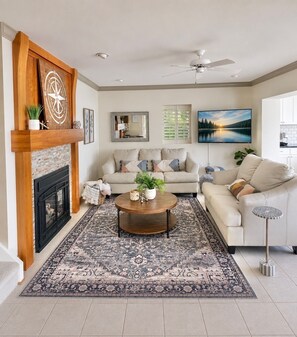 A living room with a fireplace, a ceiling fan, a large window with a view, and a coffee table. - The Lake House- FALL STAY-Cozy 3 Bedroom Lake Escape. Unwind by the Water! (Canandaigua)