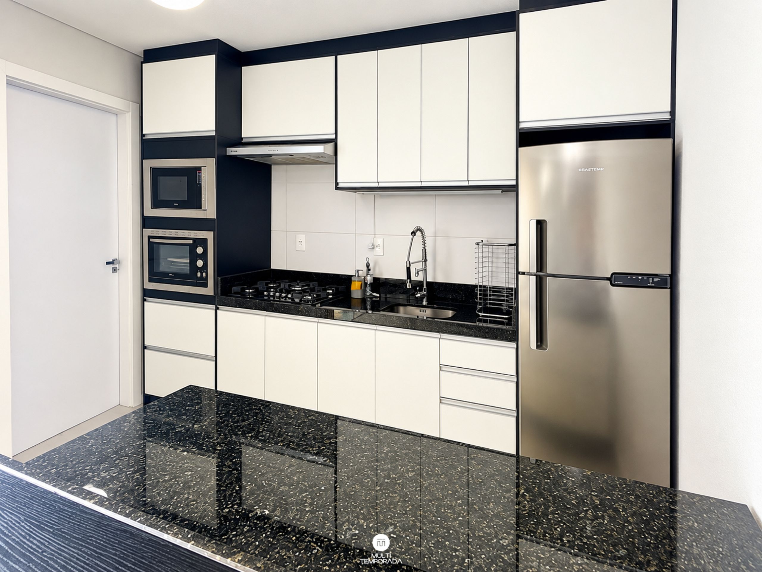 Cozinha moderna com eletrodomésticos de aço inoxidável, armários brancos e bancada de granito preto.