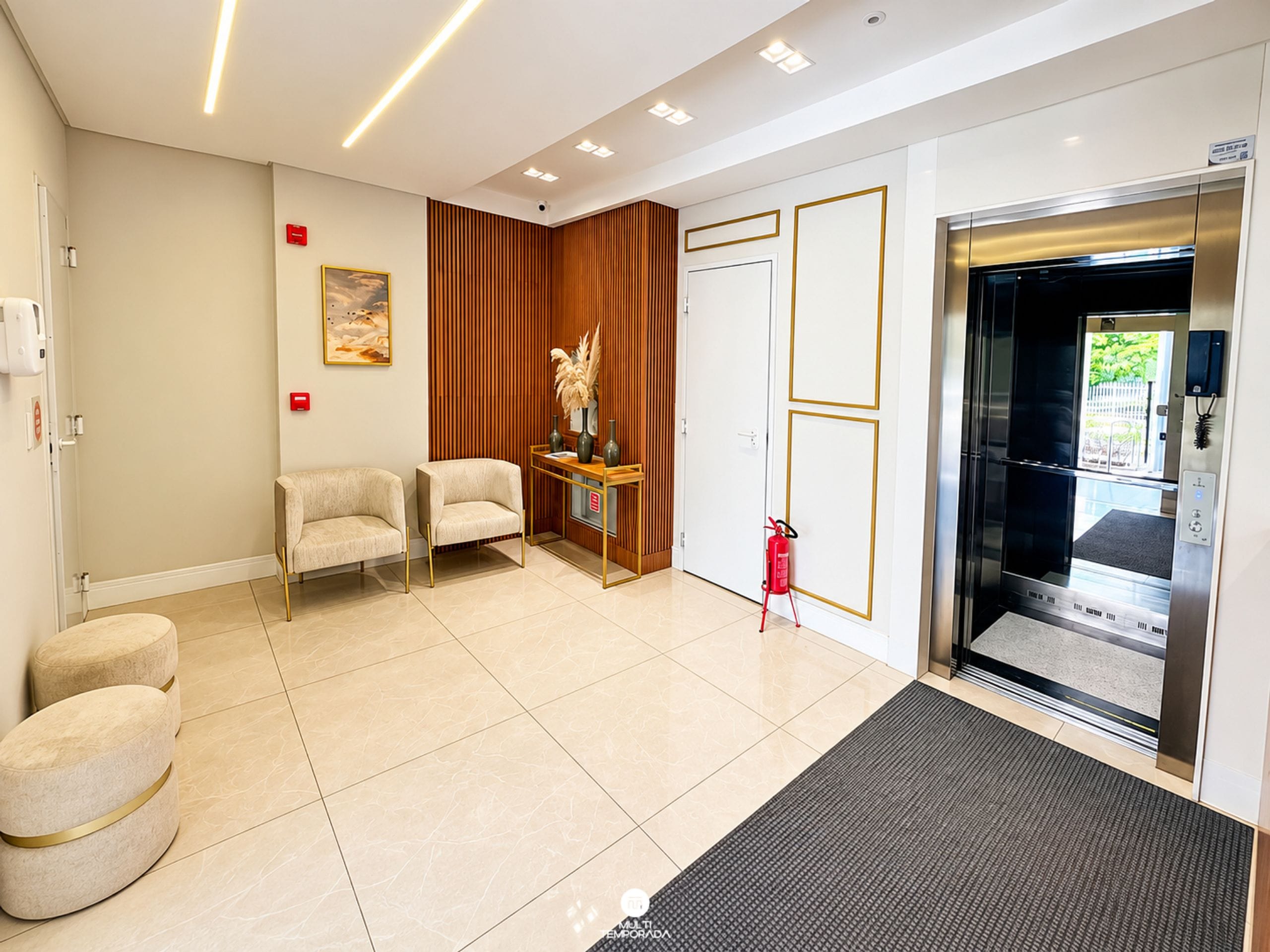 A hotel lobby with a reception desk, two armchairs, a fire extinguisher, and an elevator.
