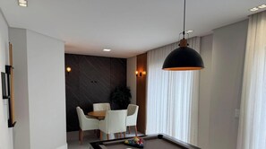 A modern living room with a pool table, a dining area with a table and chairs, and a large window with curtains. - Apartamento Infinity Residence - PB (Porto Belo)
