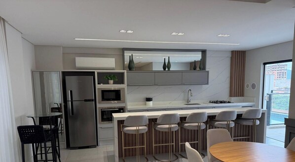 A modern kitchen with a central island, bar stools, a refrigerator, and a microwave. - Apartamento Infinity Residence - PB (Porto Belo)