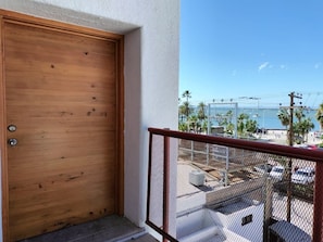 Een houten deur, een balkon met metalen hekwerk, en uitzicht op een parkeerplaats met palmbomen en een waterpartij.