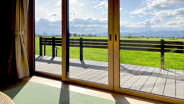 Une pièce dotée d’une grande porte-fenêtre coulissante qui donne sur une terrasse extérieure, elle-même bordée d’une clôture en bois et donnant sur un terrain herbeux.