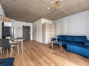 Ein moderner Wohnbereich mit blauem Sofa, einem Esstisch mit Stühlen, einer Küchenzeile mit Kühlschrank und einer Decke mit freiliegenden Holzbalken und Leuchten.