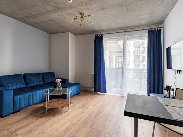 Ein modernes Wohnzimmer mit einem blauen Sofa, einem Glastisch, einem Esstisch und einem großen Fenster mit Vorhängen.