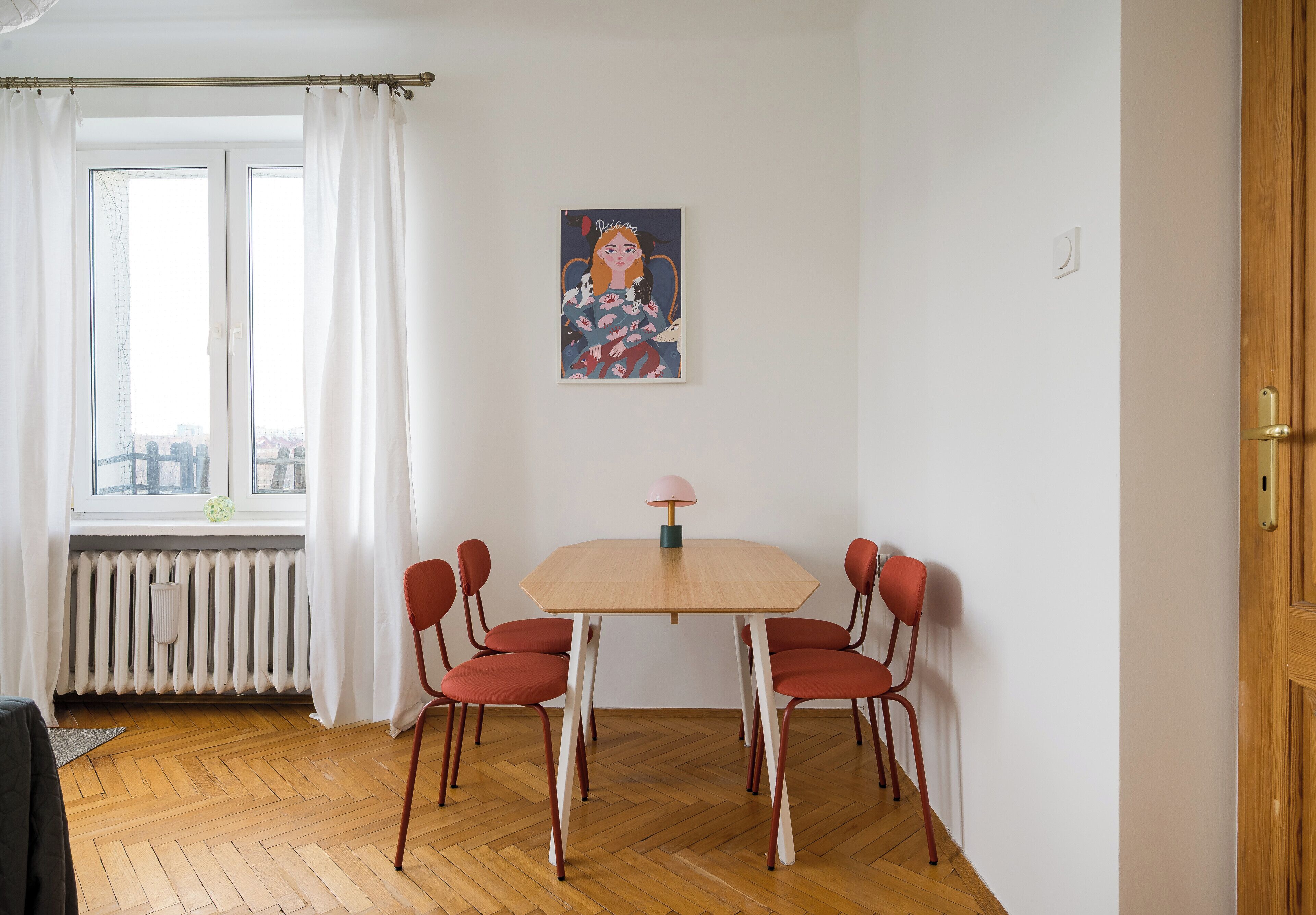 A dining area with a table and chairs, a radiator, a window with curtains, and a painting on the wall.