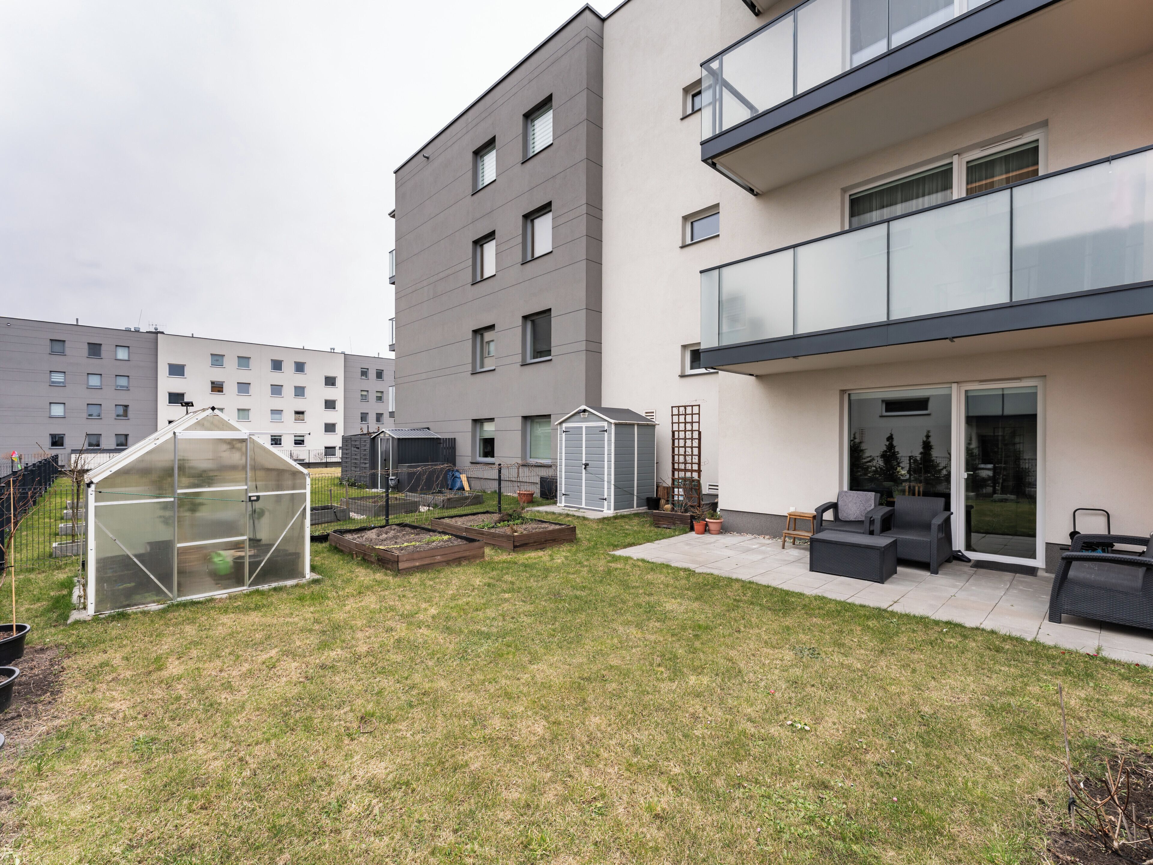 A modern building with a flat roof, a balcony with outdoor seating, and a small garden area with a greenhouse and raised garden beds.