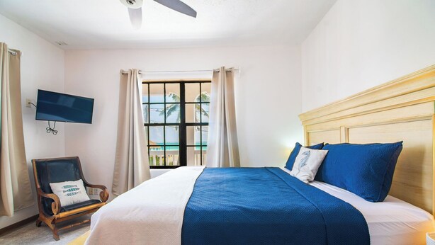 Un dormitorio con cama, una silla, un televisor y una ventana con cortinas.
