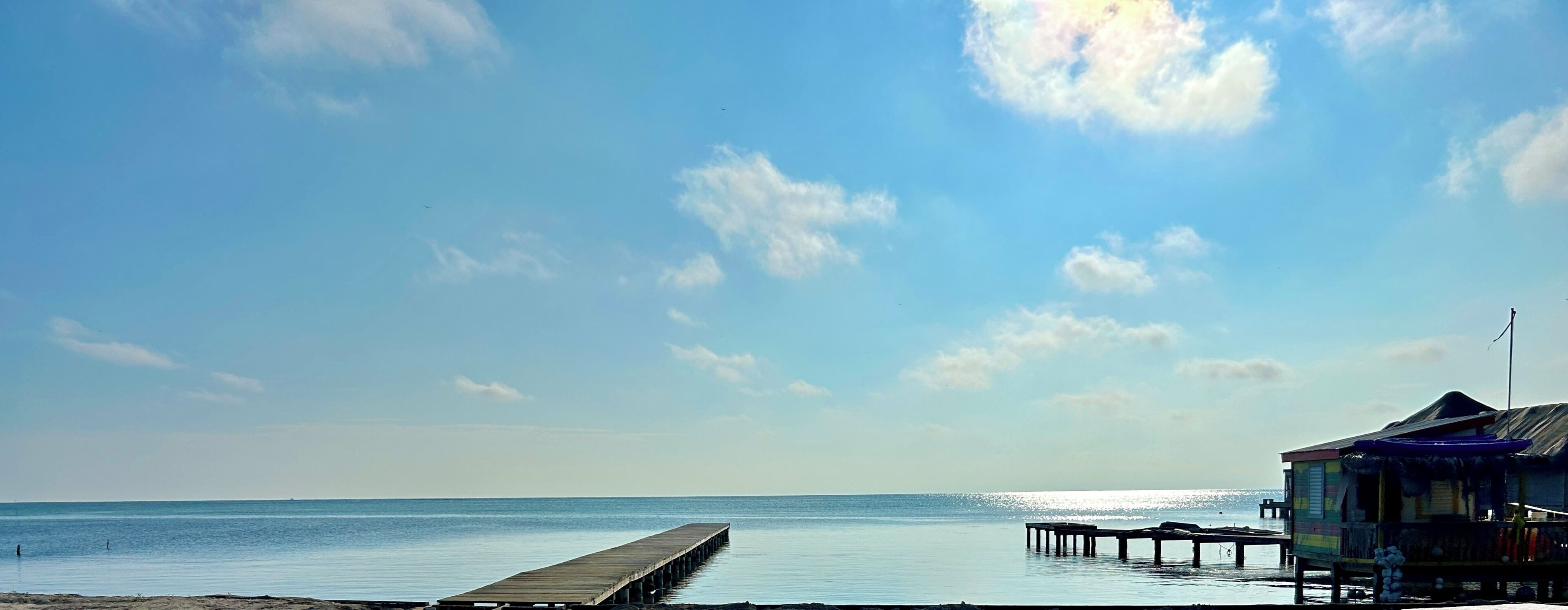 Une plage avec une jetée en bois s’avançant dans des eaux calmes, une petite cabane à toit de chaume et un ciel dégagé parsemé de quelques nuages.