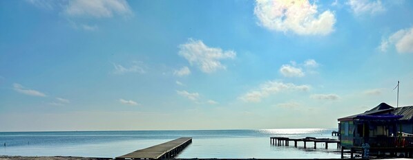 A beach with a wooden pier extending into calm waters, a small hut with a thatched roof, and a clear sky with scattered clouds.