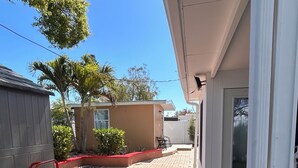 Un espace extérieur aménagé avec un sol pavé, une terrasse carrelée aux tons rouge et beige, un chariot en métal rouge et un bâtiment blanc doté d’une porte et d’une fenêtre.