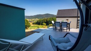 Een terras met een tafel en stoelen, een ligstoel en uitzicht op een dorp en heuvels.