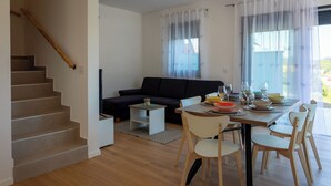 A modern living room with a staircase, a black sofa, a dining table set for four, and a view through large windows. - Modern vineyard home with private jacuzzi (Margečan)