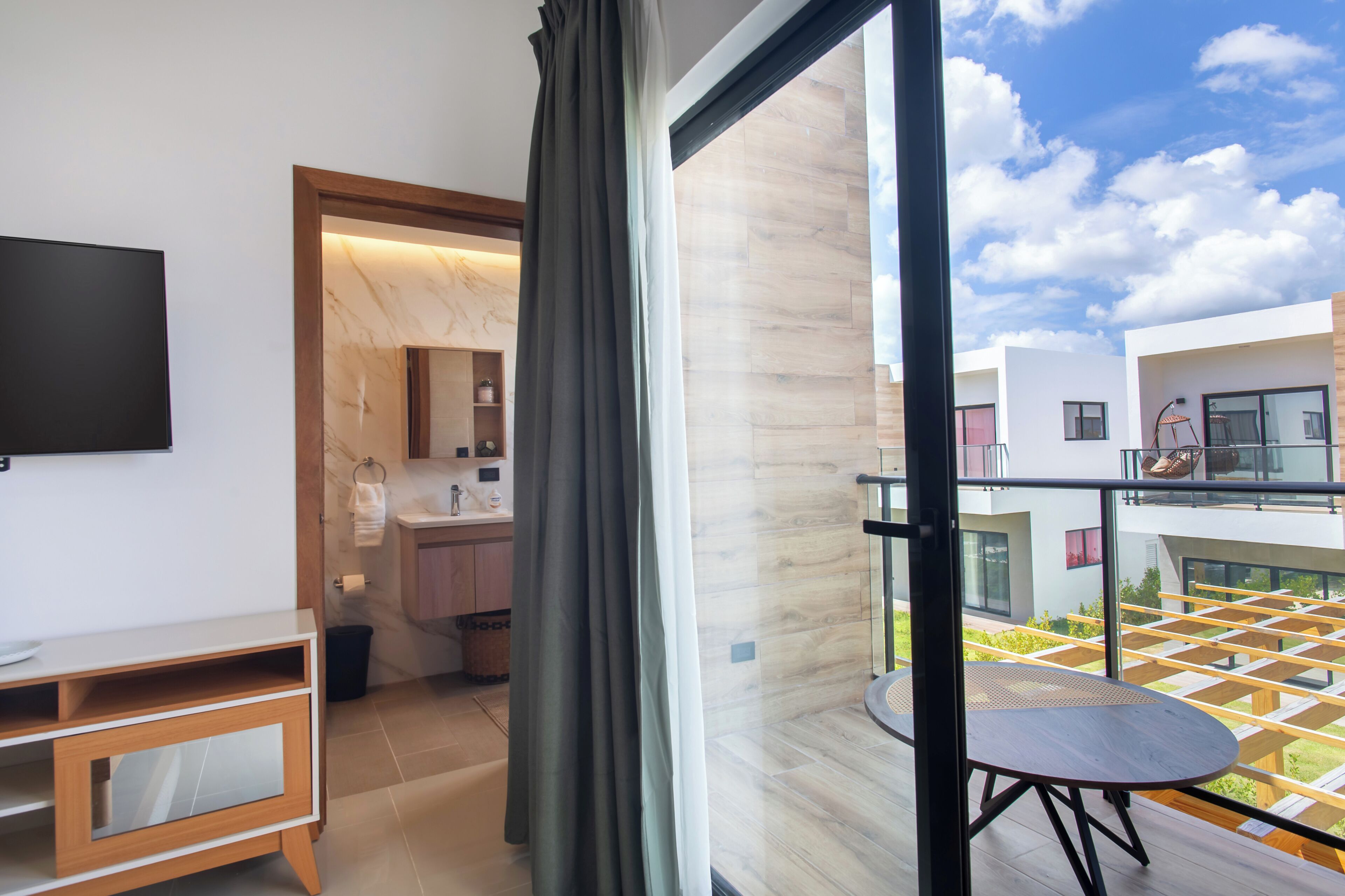 A modern room with a TV, a wooden cabinet, a balcony with a table, and a view of the sky.