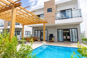 Een modern huis met twee verdiepingen, een houten pergola, een zwembad en een terras met tuinmeubilair.