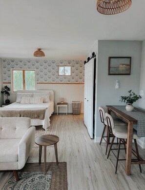 A bedroom with a bed, bedside tables, a desk, chairs, a hanging light fixture, and a window with a view.