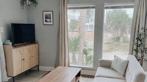 A living room with a sofa, a coffee table, a TV on a cabinet, and large windows with curtains. - Pet Friendly Studio / Walk to Beach & Eats (Panama City Beach)