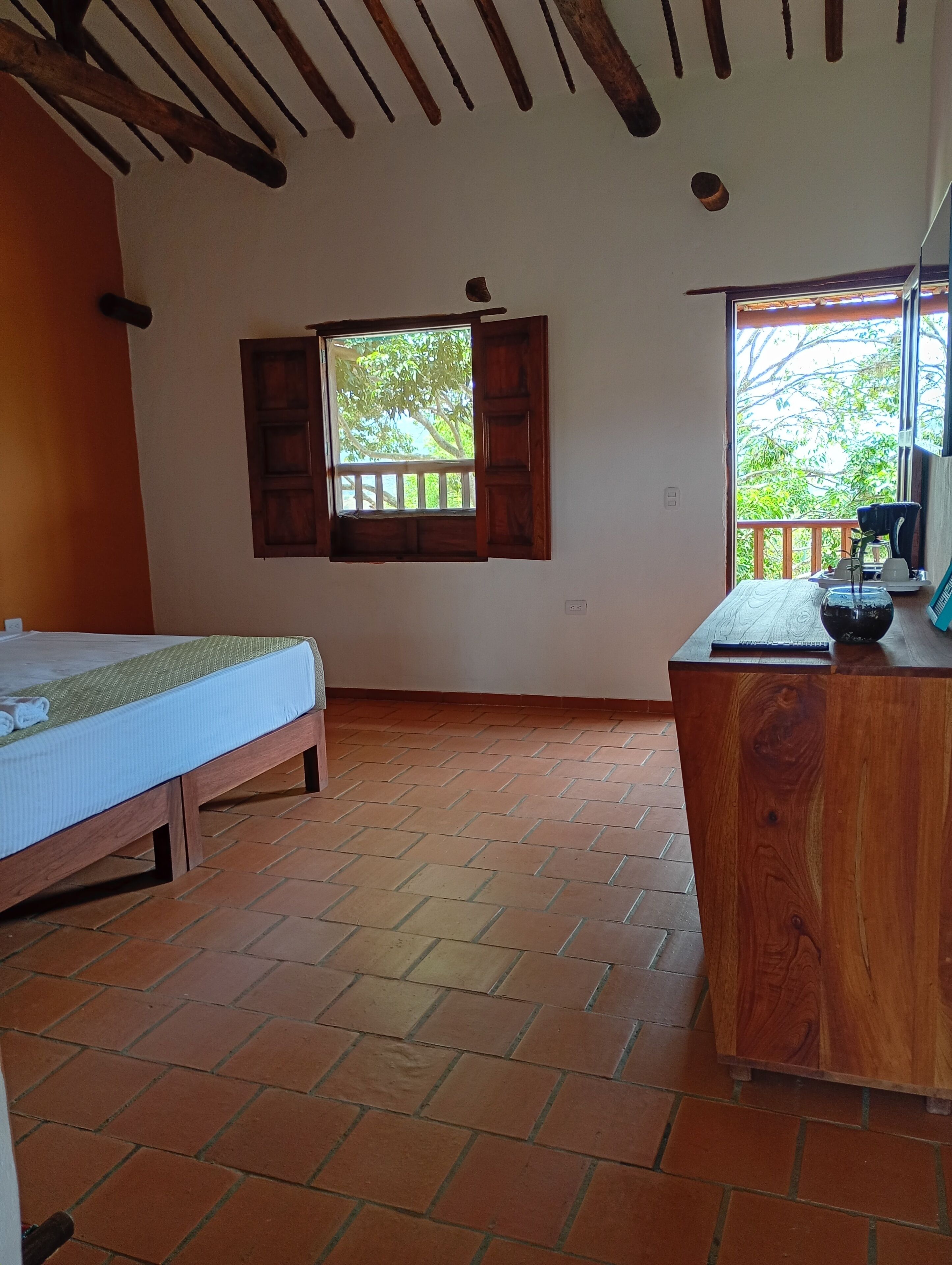 Um quarto com cama, escrivaninha de madeira, janela com persianas abertas e piso ladrilhado.