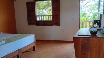 Um quarto com cama, escrivaninha de madeira, janela com persianas abertas e piso ladrilhado.