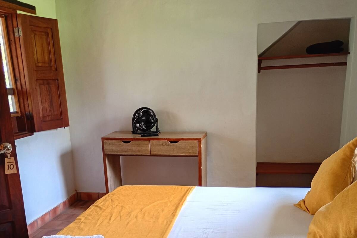 A bed with a white mattress and yellow pillows, a wooden bedside table with a fan, and a wooden wardrobe.