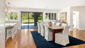 Un vaste espace dédié à la salle à manger, avec une grande table et des chaises blanches, une cuisine dotée d’un bar et une porte-fenêtre coulissante donnant sur un jardin.