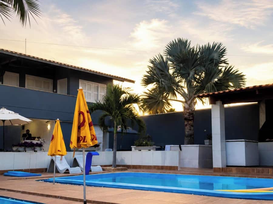 Une piscine avec des chaises longues et des parasols, un bâtiment moderne doté d’un toit plat, et des palmiers.