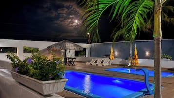 Un área de piscina en la azotea con dos piscinas, sillas de descanso, una sombrilla de paja y una palmera.
