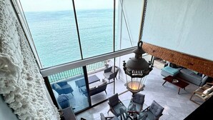 Et rum med højt til loftet og et stort vindue med udsigt over havet, møbleret med moderne stole og et bord, samt en hængende lygte.