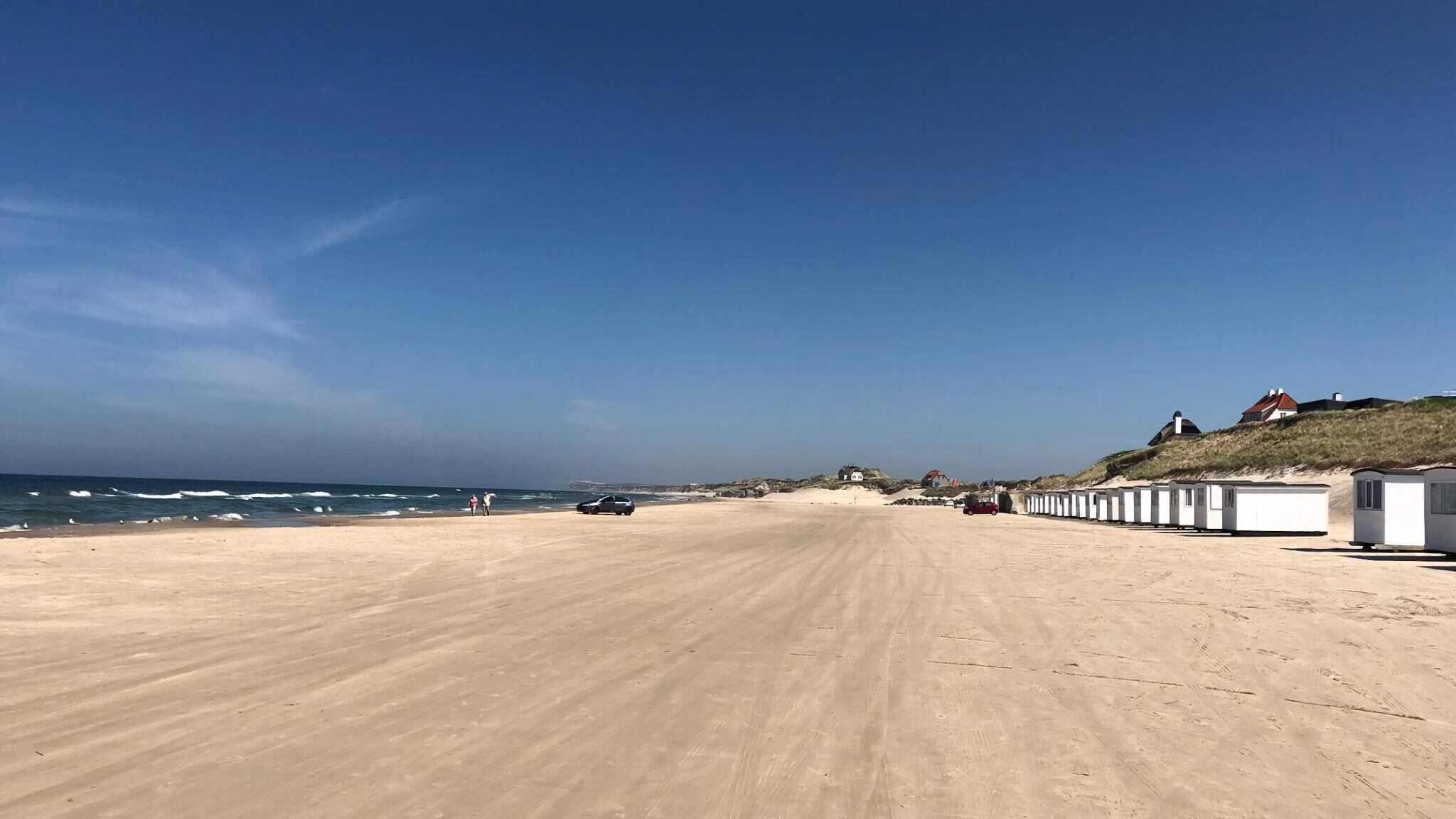 Una playa arenosa con una hilera de cabañas blancas de playa, algunas personas y un coche estacionado en la arena.