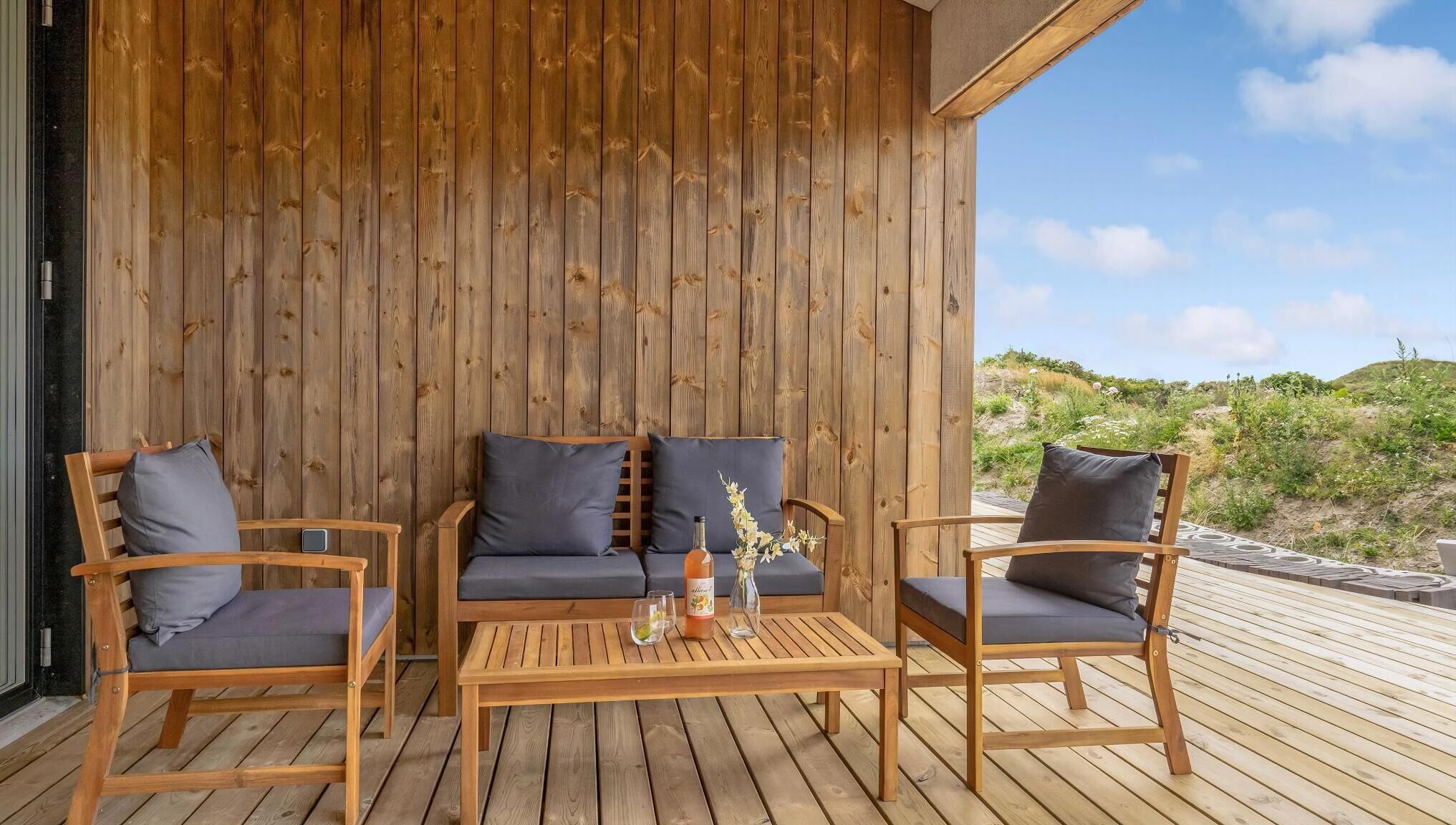 En træterrasse med sofa og stole, et bord med en flaske og glas, og udsigt over landskabet.