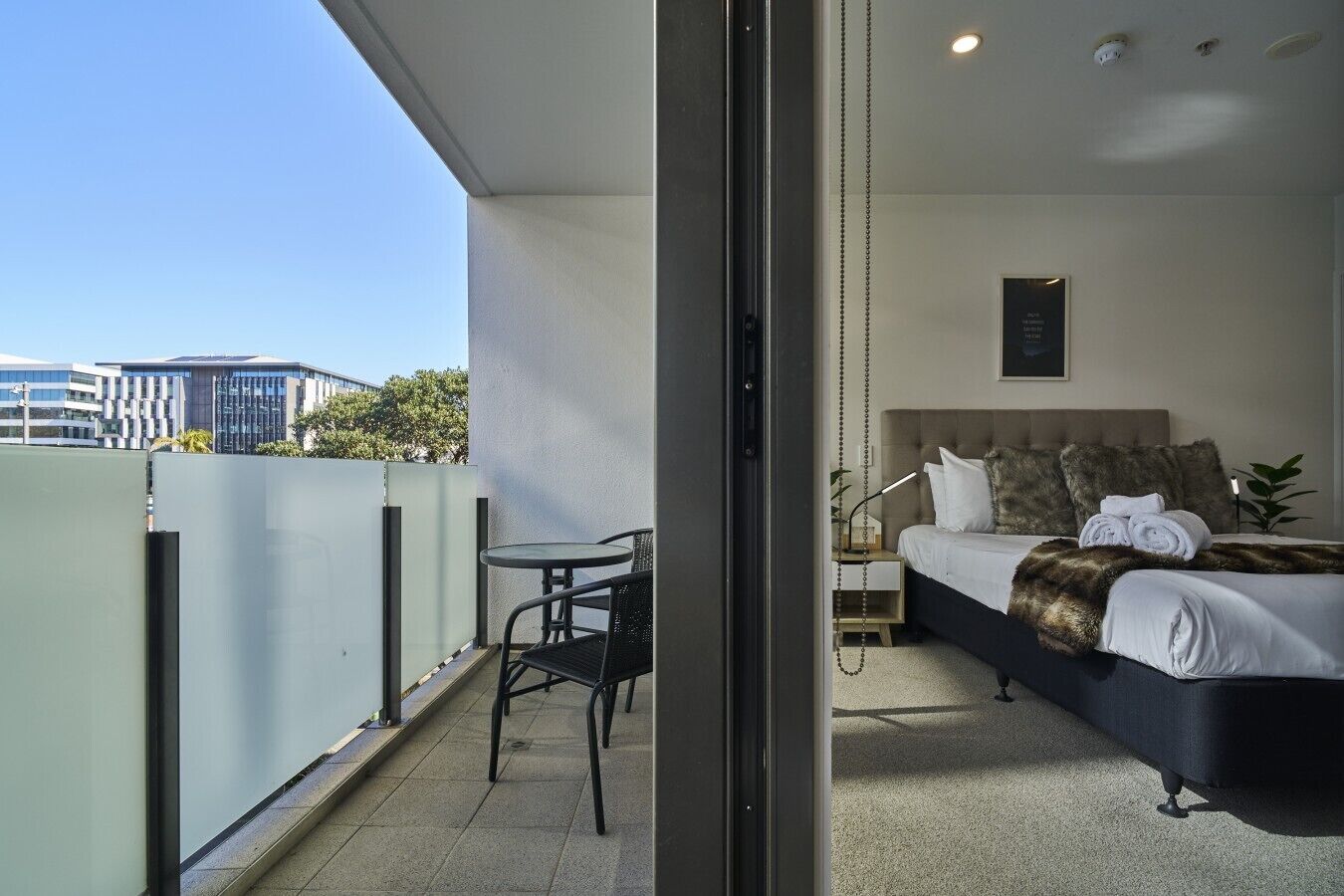 A balcony with a table and chairs, a bed with a headboard, and a view of buildings in the background.