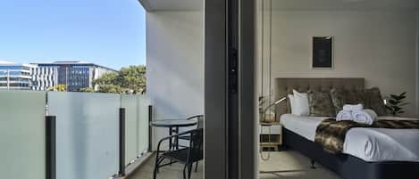 A balcony with a table and chairs, a bed with a headboard, and a view of buildings in the background.