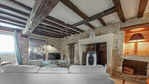 Een rustieke kamer met zichtbare balken, een stenen muur, een open haard, een televisie en een bank.