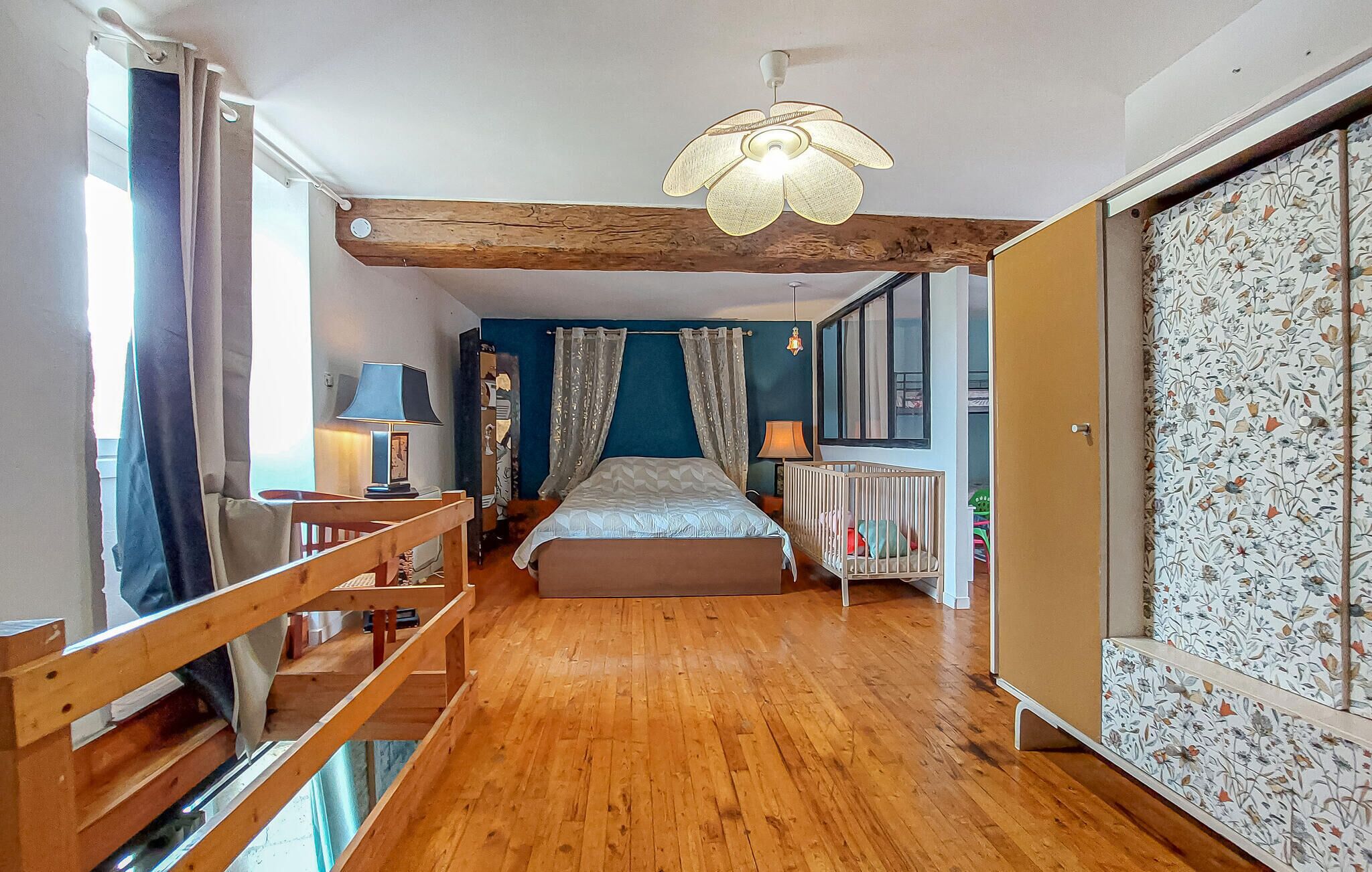 A bedroom with a wooden floor, a bed, a crib, a ceiling fan, and a window with curtains.
