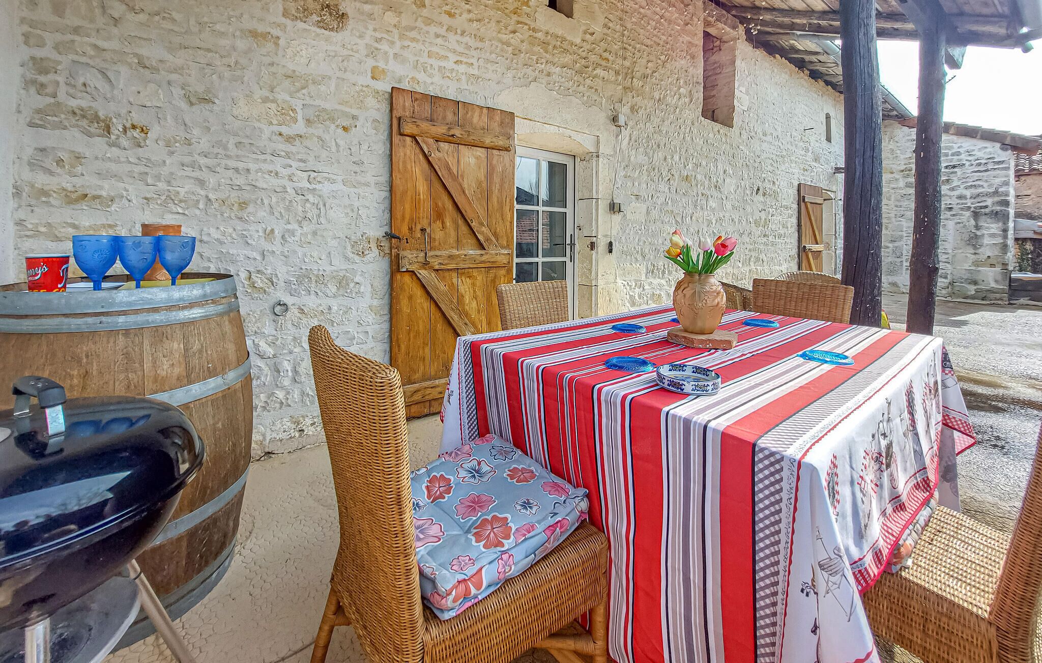 A rustic outdoor dining area with a table set for two, a wicker chair, a wooden barrel, and a rustic wall.