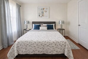 A bedroom with a bed, two bedside tables, a nightstand, a door, and a window with curtains.