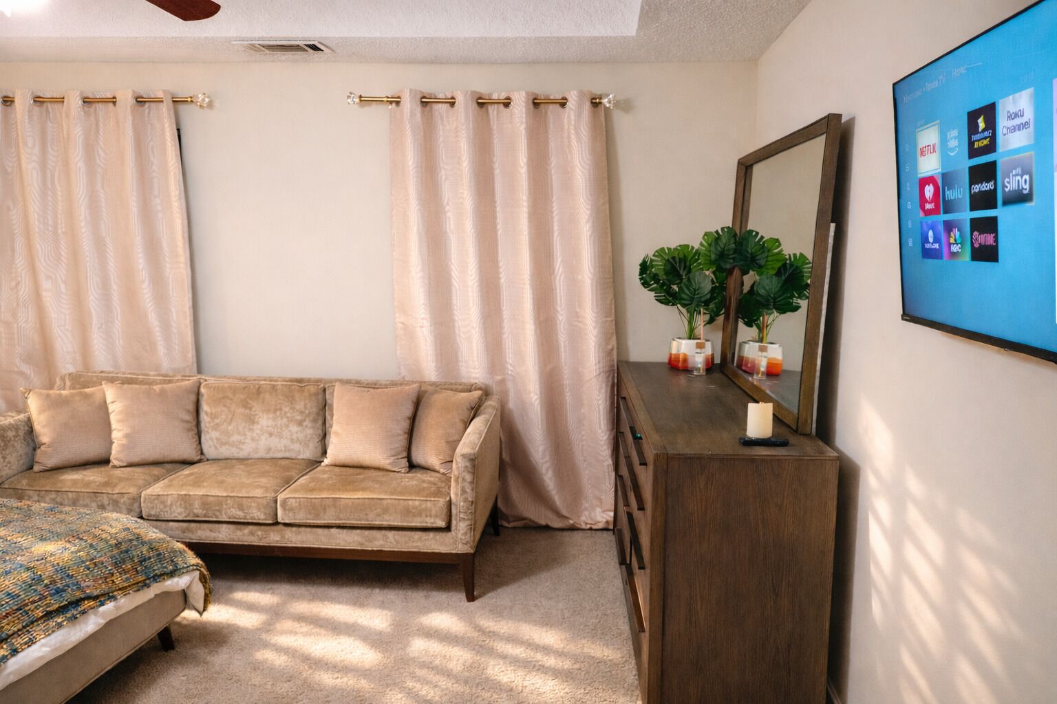 Quarto com sofá bege, cómoda de madeira, cama com manta estampada e televisão na parede exibindo serviços de streaming.