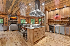 A wooden interior with a kitchen island, bar stools, and a stone fireplace. - Luxury Riverfront Retreat • Spa • Game Room • Minutes to Downtown Blue Ridge (Blue Ridge)