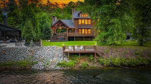 A rustic cabin with a stone chimney, a deck with chairs, and a river in the foreground. - Luxury Riverfront Retreat • Spa • Game Room • Minutes to Downtown Blue Ridge (Blue Ridge)