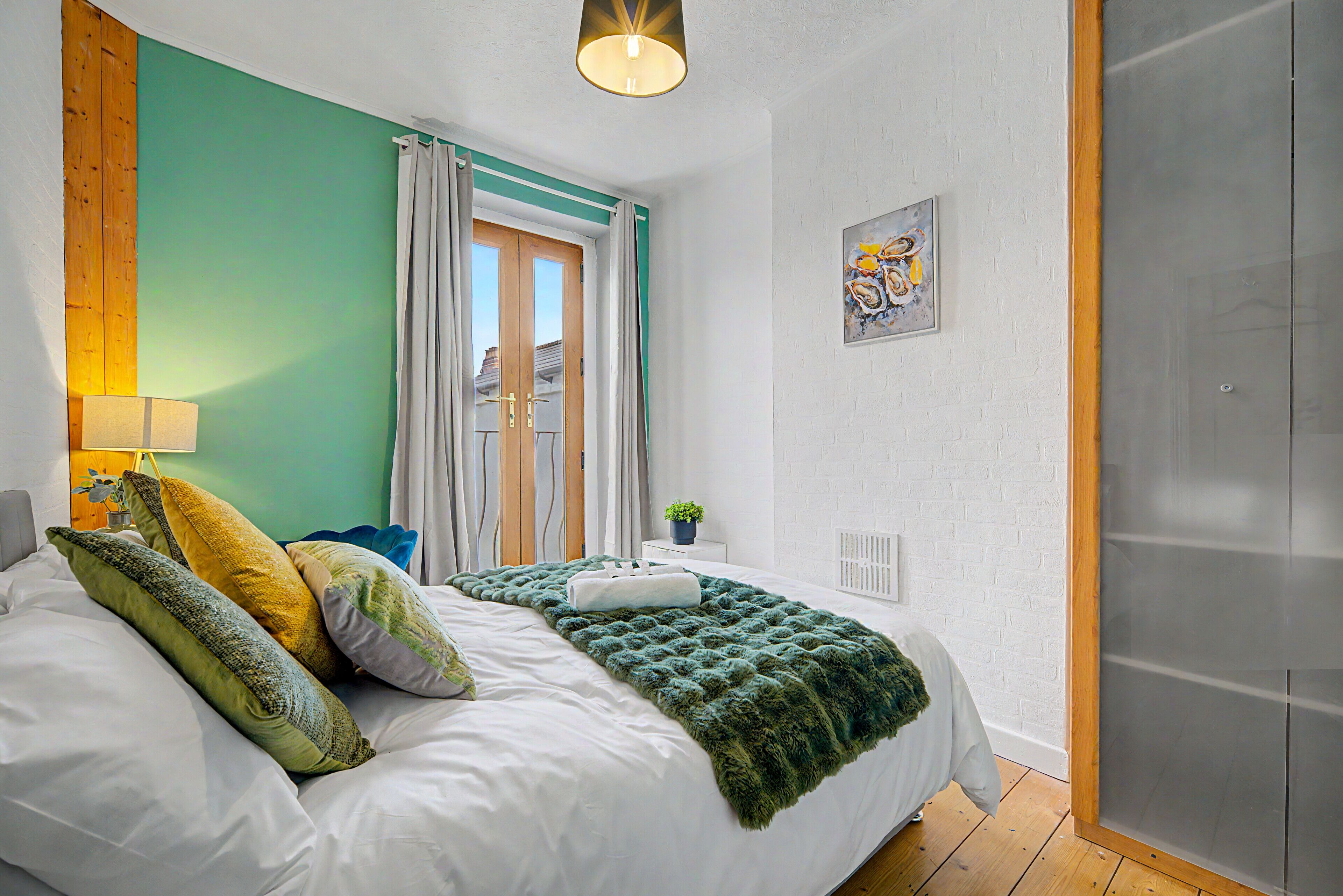 Un dormitorio con cama, almohadas, manta, lámpara, ventana con cortinas, planta y puerta.
