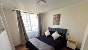 A bedroom with a bed, a nightstand, a window with curtains, and a painting on the wall. - Cozy apartment in front of Torpederas beach, ideal for resting. (Valparaíso)