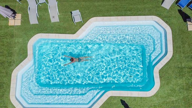 Piscina rectangular con una persona nadando, rodeada de césped y sillas de descanso.
